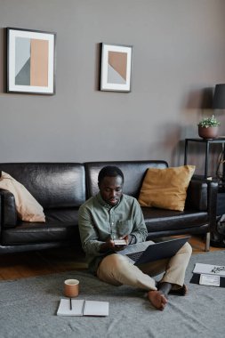 Genç Afrikalı Amerikalı adamın oturma odasında oturup dizüstü bilgisayar ve akıllı telefon kullanarak finans stratejisi üzerinde çalışmasının dikey çekimi.