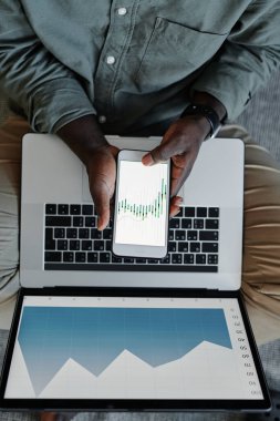 Yukarıdan bakıldığında, genç siyahi bir adamın dizüstü bilgisayar ve akıllı telefon kullanarak finans projesi üzerinde çalıştığı görülüyor.