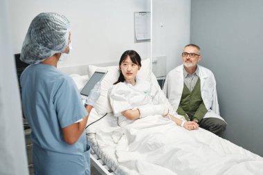 Tanımlanamayan doktor elinde dijital tabletle acil serviste genç Asyalı hasta ve akrabasıyla konuşuyor.