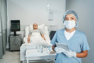 Koruyucu maske takan bir doktorun yatay portresi ve dijital tablet tutan eldivenler, kameraya bakan hasta arka planda yatağında yatıyor.