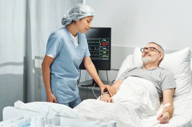 Genç, Latin asıllı bir doktor, hastane odasındaki HRM cihazıyla yetişkin bir erkek hastanın kalp atışlarını izliyor.