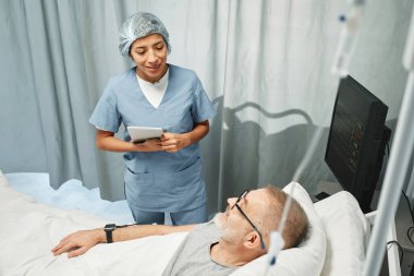 Hastanede elinde dijital tabletle çalışan yetişkin bir kadının olgun bir erkeğe yatakta bir şeyler söylediği yatay açılı bir fotoğraf.