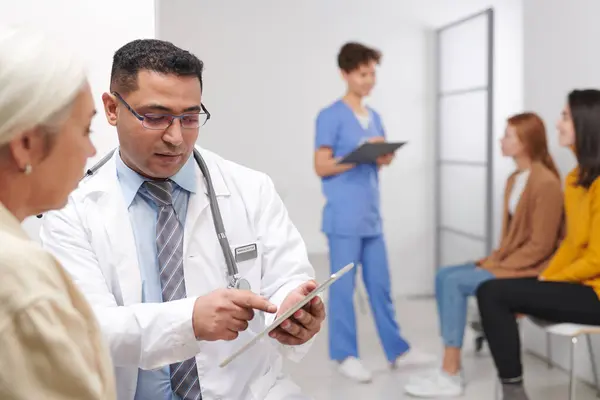 Middle-Eastern doctor showing test results to woman while his colleague calling for next patient in queue