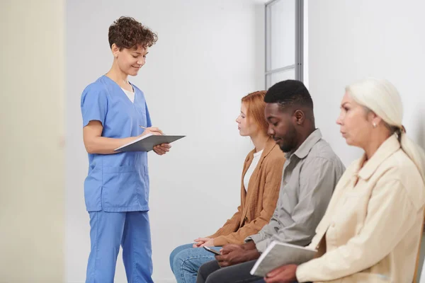 Group of three multi-ethnic people sitting in queue waiting for consultation in modern hospital, medical worker standing near checking attendance