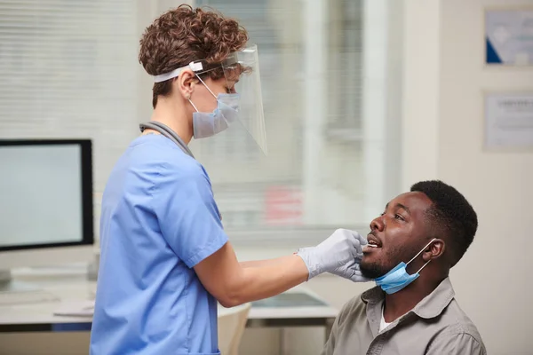 Horizontal side view shot of doctor wearing protective masks testing African American patient for viral disease