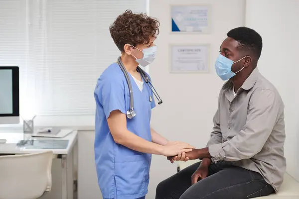 Horizontal side view medium long shot of female doctor wearing mask on face comforting African American male patient holding his hand