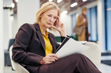 Ciddi bir kadın avukat, adliyenin lobisinde otururken belgeleri okuyor ve toplantı için hazırlanıyor.