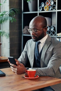 İnternetteki işine yoğunlaşan Afrikalı işadamı cep telefonuyla masada oturuyor ve kafede kahve içiyor.