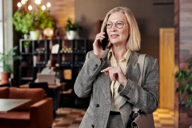 Gözlüklü ve şık takım elbiseli olgun bir iş kadını iş görüşmesi için restorana gelirken cep telefonuyla konuşuyor.