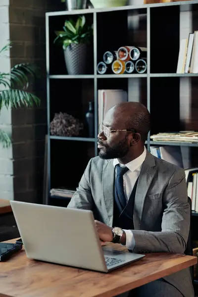 African man in suit looking through the window with pensive sight while working online on laptop in cafe