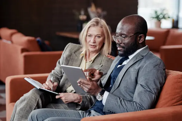 African businessman pointing at the screen of tablet pc with businesswoman making notes in notebook, they planning work together sitting in cafe