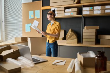 Dijital tablet kullanan genç bir kadın kargo için paket dağıtıyor, depoda çalışıyor.