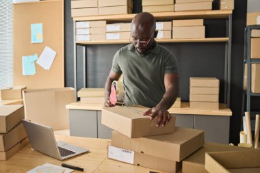 Afrikalı genç işçi karton kutular hazırlıyor ve ambalajda yapışkan bantlarla paketleniyor.