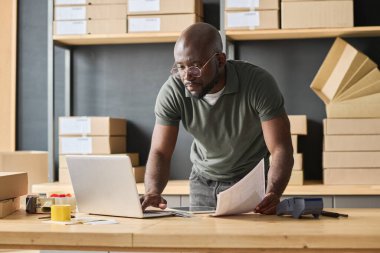 Ciddi bir Afrikalı adam masadaki dizüstü bilgisayarına odaklanmış, depolama odasında çalışırken paketlerin verilerini yazıyor.