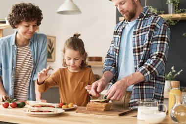 Küçük çocuk, anne ve babasına evcil mutfaktaki masada sebzeli sandviç pişirmeleri için yardım ediyor.