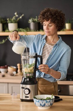 Mutfaktaki masada meyve suyu hazırlamak için blender 'a süt ekleyen genç bir kadın.