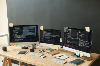 Ofisteki modern bilişim mühendisinin çalışma yerinde duran ekranlarında şifreli veri ve yapışkan not kağıtları olan üç bilgisayar monitörü.