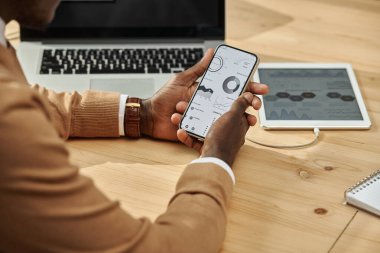 Afrika kökenli Amerikalı iş adamının, bilgisayar aygıtıyla işyerinde otururken finansal grafiklerle çalışmak için akıllı telefon kullandığı yakın plan.