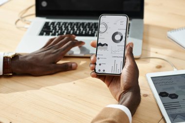 Bilgisayarlı masada otururken akıllı telefonunda finansal grafiklerle online çalışan Afrikalı Amerikalı adamın yakın plan fotoğrafları.