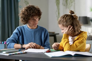 Young teacher teaching difficult child at home, they sitting together at desk with books and talking clipart