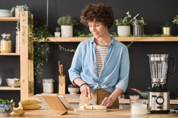 Young woman with curly hair cutting banana on cutting board at table and watching video on digital tablet