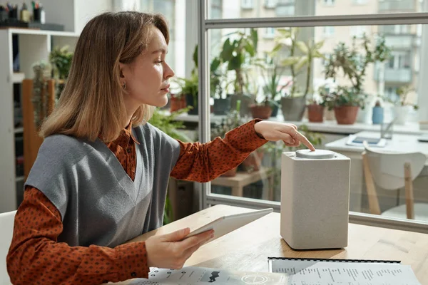 Young woman connecting smart speaker with tablet pc while working at modern office with green plants