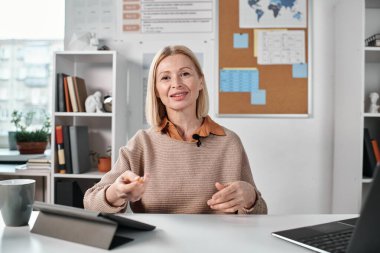 Sınıfta masada oturmuş öğrencilere online ders verirken kameraya bakan olgun öğretmenin portresi