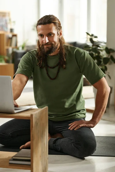 Yoga instructor sitting on the floor in front of laptop and talking to his students online