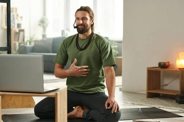 Young yoga teacher sitting on the floor in front of laptop and talking to his students online during yoga lessons
