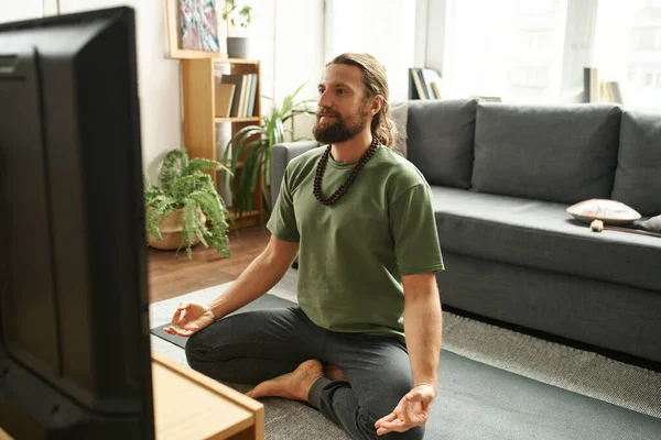 Bearded man sitting on the floor on exercise mat in front of TV and watching it while doing yoga in living room