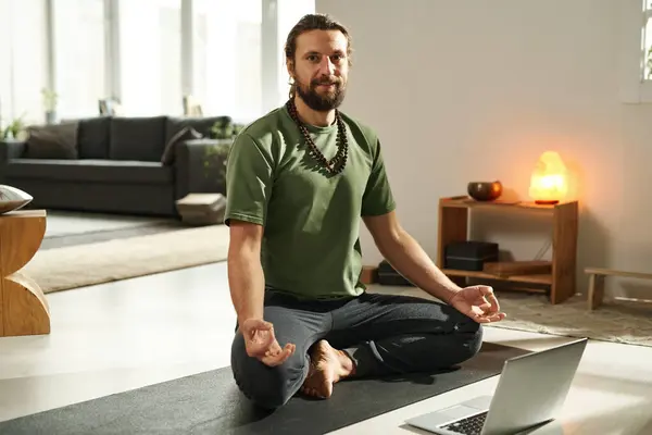 Portrait of young man meditating in lotus position on exercise mat at home, he learning to do yoga online on laptop