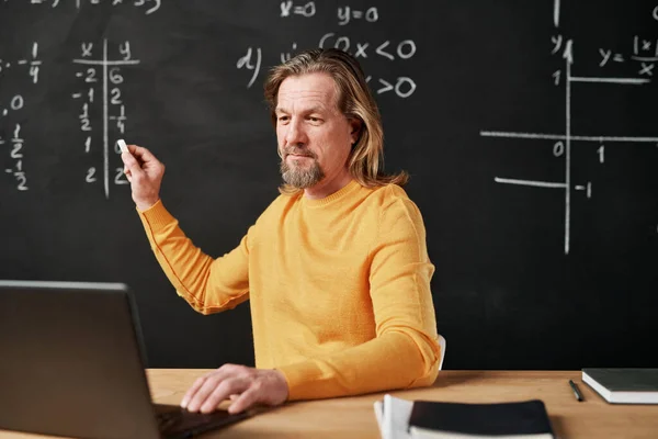 Mature bearded teacher writing on blackboard and using laptop for online lesson in the classroom