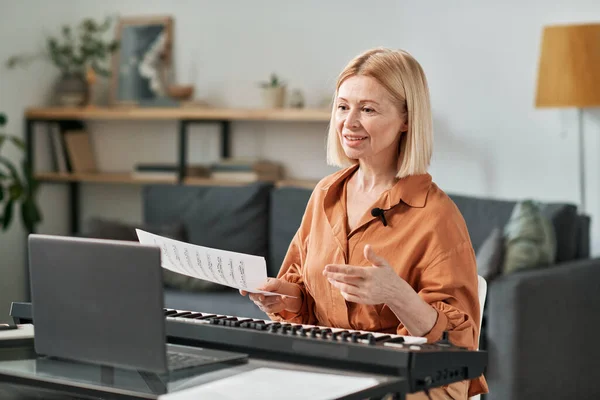 Mature woman sitting at table in front of laptop and teaching musical lesson online