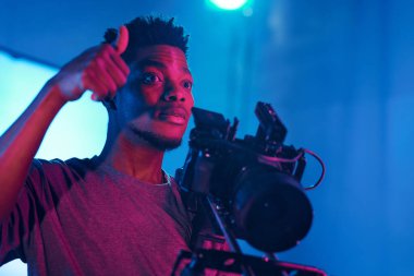 Yapım stüdyosunun operatörü çekim sırasında kameranın önünde dikilirken ekibine başparmağını gösteriyor.