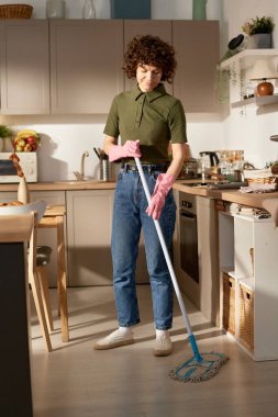 Evdeki mutfakta paspasla yerleri temizleyen genç kadının dikey görüntüsü.