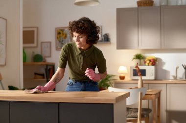 Ev hanımı kulaklık takıp müzik dinliyor. Mutfakta ev işi yaparken masayı paçavrayla siliyor.