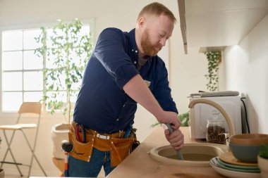 Sakallı tamirci mutfaktaki tıkanıklığı kaldırmak için bir araç kullanıyor.