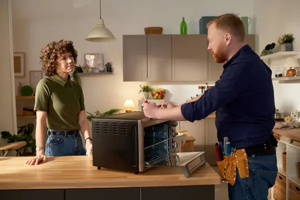 Professional serviceman repairing broken microwave oven and talking to housewife while they standing in the kitchen