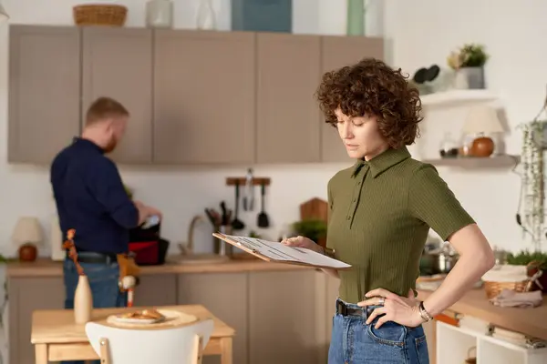 Housewife examining customer service contract standing in the kitchen with repairman working in the background