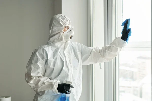Cleaning worker in protective suit wiping windows with rag and detergent during work in apartment