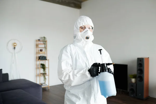 Portrait of cleaning worker in protective suit looking at camera while disinfecting apartment with detergent