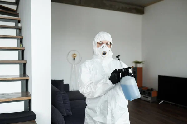 Portrait of cleaning worker in protective suit looking at camera while spraying detergent with sprayer during housework