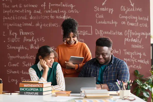 Front view portrait of three Black college students looking at laptop screen against blackboard with writings in background