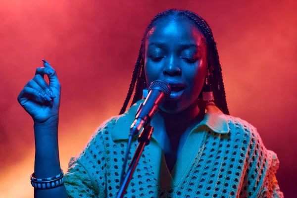 Front view portrait of young Black woman singing into microphone performing on stage in neon lights