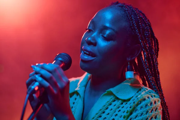 Close up portrait of young African American woman with braided hair singing into microphone in red and blue neon lights