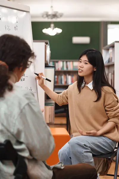 Vertical portrait of young Asian woman explaining grammar rules of Chinese language to students in library and pointing at board