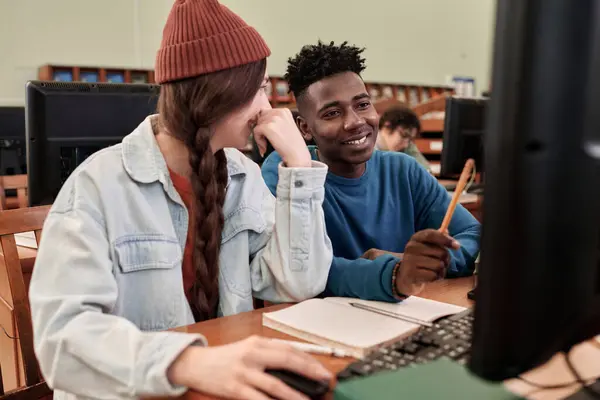 Portrait of two young students using computer in college library and pointing at screen while working on project together