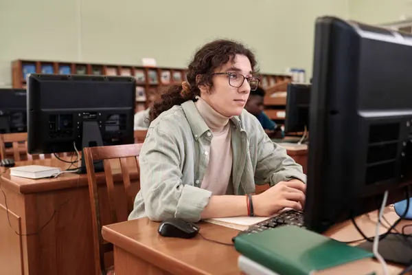Portrait of long haired male student looking at computer screen doing online studying in college library copy space