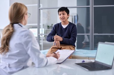 Klinikteki çocuk doktoruna el ve bilek ağrısı belirtilerini açıklayan genç bir çocuğun portresi.