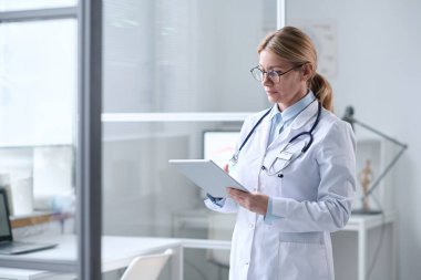 Sarışın, yetişkin bir kadının portresini panoya yazıp temiz beyaz ofis fotokopi alanında duran bir doktor olarak göster.
